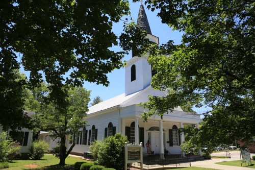 A white church with a tall steeple, surrounded by green trees and shrubs on a sunny day.