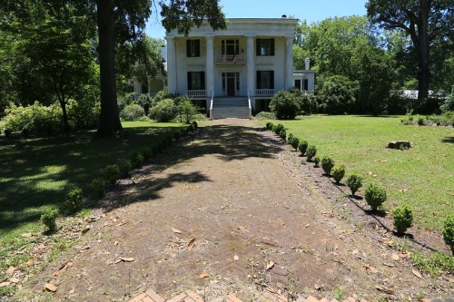 A grand, two-story mansion with white columns, surrounded by a manicured lawn and a pathway lined with small shrubs.