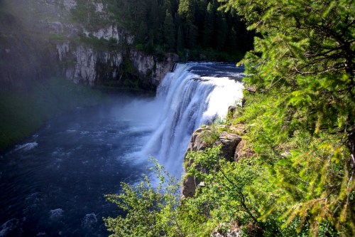 A stunning waterfall cascades over rocky cliffs, surrounded by lush greenery and a serene river below.
