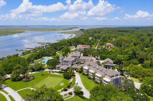 Aerial view of a large estate surrounded by lush greenery and water, featuring a pool and expansive grounds.