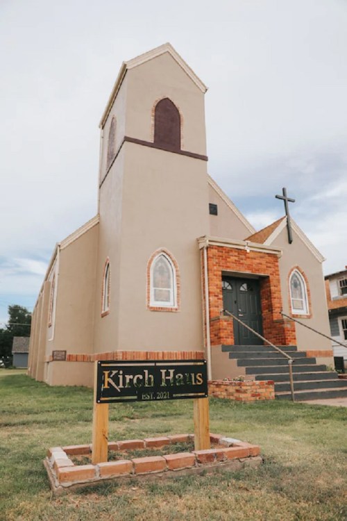 A beige church building with a cross, featuring a sign that reads "Kirch Haus, EST. 2021," set against a cloudy sky.