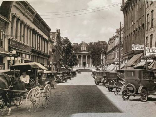 Historic street scene featuring horse-drawn carriages and early automobiles, with a gazebo in the background.
