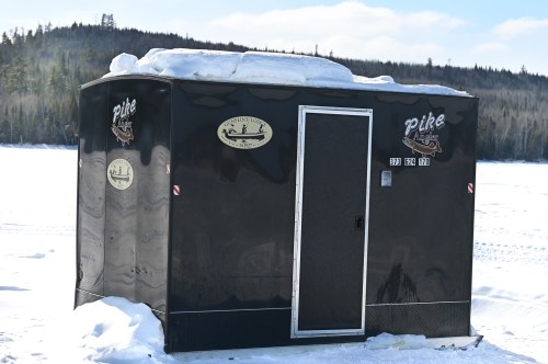 A black ice fishing shelter with snow on top, set against a snowy landscape and trees in the background.