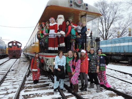 A festive group in holiday attire poses with Santa on a train car in a snowy setting.
