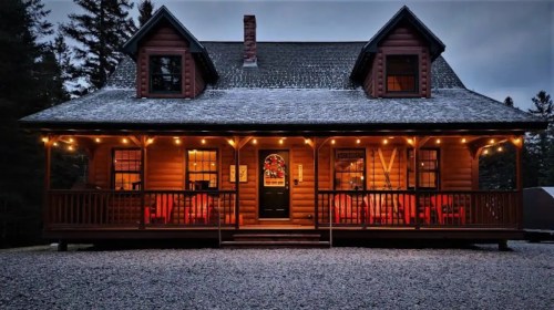 A cozy log cabin with a porch, decorated with lights, surrounded by trees at dusk.