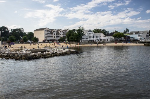 A sandy beach with people enjoying the sun, surrounded by water and buildings under a partly cloudy sky.