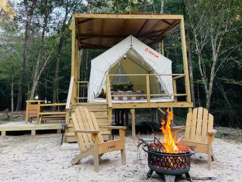 A cozy glamping tent on a wooden platform, surrounded by trees, with a fire pit and wooden chairs in front.