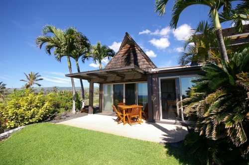 A tropical house with a wooden deck, surrounded by palm trees and lush greenery, under a clear blue sky.