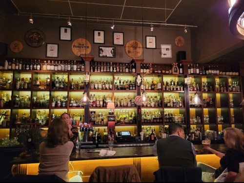 A cozy bar with a wooden shelf filled with various bottles, dim lighting, and patrons enjoying drinks at the counter.