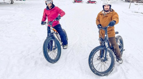 Two men on fat bikes in the snow, smiling and wearing helmets, with snowmobiles in the background.