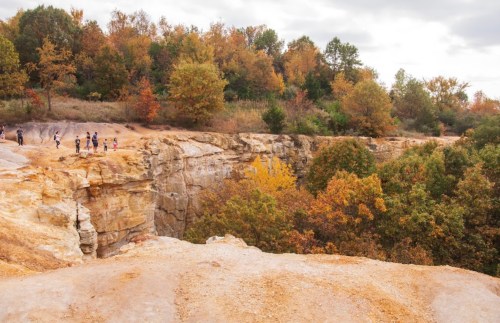 A rocky cliff with people walking along the edge, surrounded by vibrant autumn foliage and a cloudy sky.