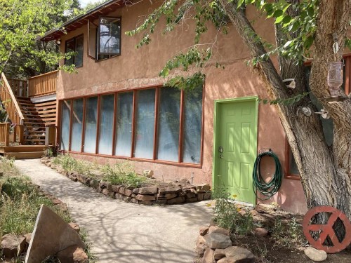 A rustic house with a green door, large windows, and a stone pathway, surrounded by trees and greenery.