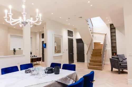 A modern dining area with a chandelier, blue chairs, and a staircase leading to an upper level.