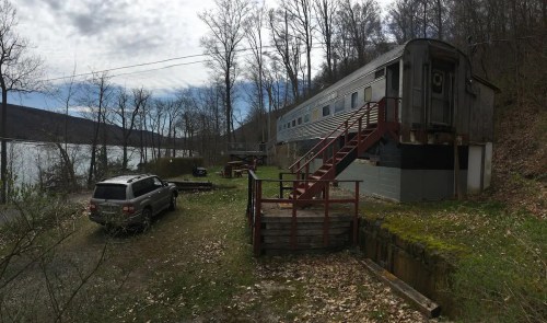 A vintage train car converted into a cabin, set by a lake with trees and a parked SUV nearby.