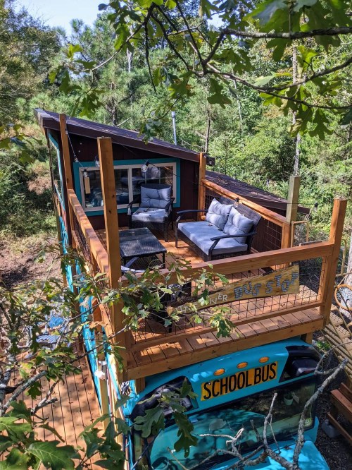 A converted school bus with a wooden deck and seating, surrounded by trees in a natural setting.