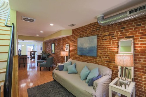 Cozy living area with a brick wall, sofa, lamps, and a staircase leading to a dining space. Bright and inviting atmosphere.
