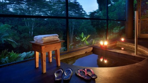 A serene spa setting with a wooden stool, towels, candles, and a view of lush greenery through large windows.