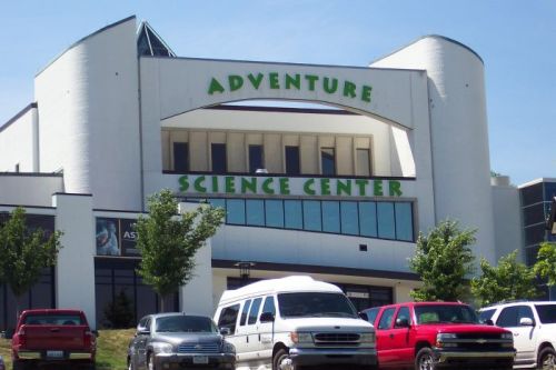 Exterior of the Adventure Science Center, featuring a modern design and green signage, with parked cars in front.