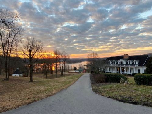 A scenic view of a driveway leading to a house, with a sunset over a lake and cloudy sky in the background.