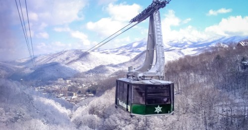 A scenic cable car glides over snow-covered mountains under a blue sky with fluffy clouds.