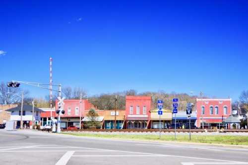 Colorful storefronts line a small town street under a clear blue sky, with traffic signs and a railroad crossing nearby.