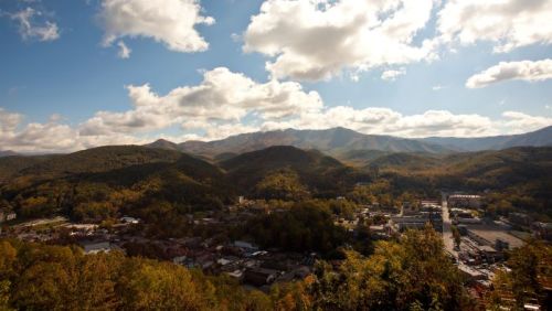 A scenic view of a mountain town surrounded by lush hills and a blue sky with fluffy clouds.