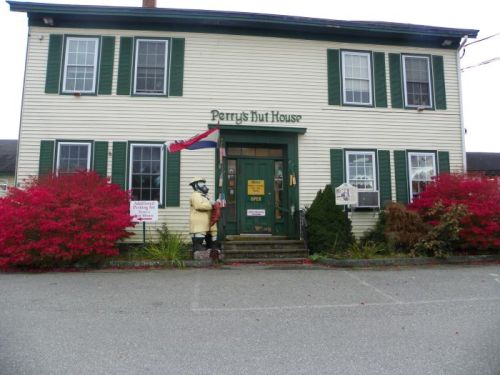 A two-story building with green shutters, a sign reading "Perry's Hut House," and vibrant red bushes in front.