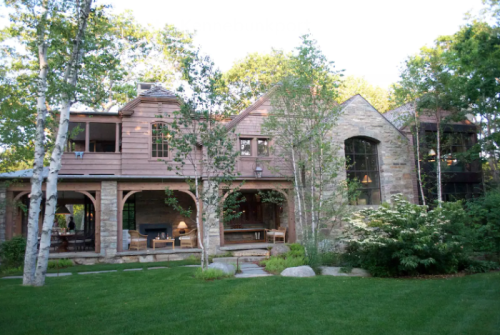 A beautiful stone and wood house surrounded by trees and a well-maintained lawn, featuring large windows and a porch.