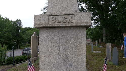 A granite gravestone with the name "BUCK" engraved, surrounded by a cemetery and American flags.