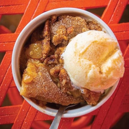 A bowl of warm dessert topped with a scoop of vanilla ice cream, featuring a crumbly, sweet topping and pecans.