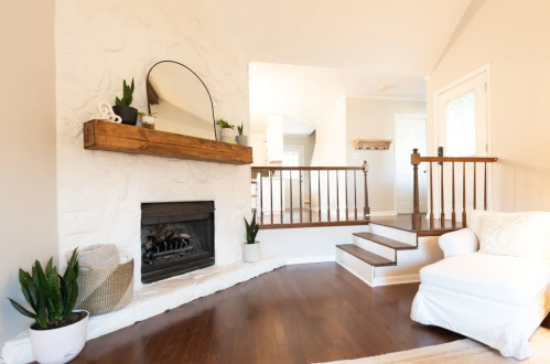 Cozy living room with a white stone fireplace, wooden mantel, plants, and a white armchair beside a staircase.