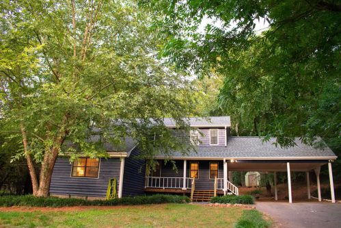 A cozy blue house surrounded by trees, featuring a front porch and a carport, set in a serene outdoor environment.
