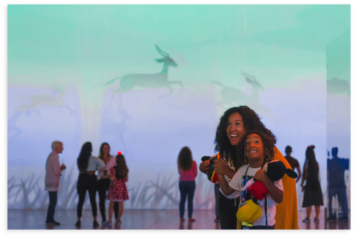 A woman and a child smile in an art installation, surrounded by colorful projections and other visitors.