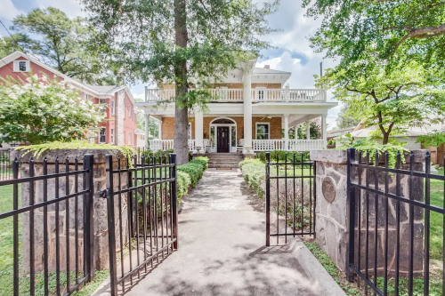 A large, two-story house with a porch, surrounded by greenery and a gated entrance.