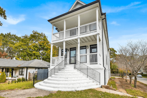 A two-story white house with a large front porch and steps, surrounded by trees and a quiet neighborhood.