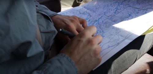 A person’s hands holding a pen, writing on a large map while seated in a vehicle.
