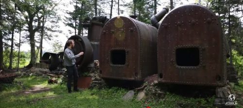 A person stands near two large, rusty industrial boilers in a forested area, surrounded by trees and remnants of machinery.