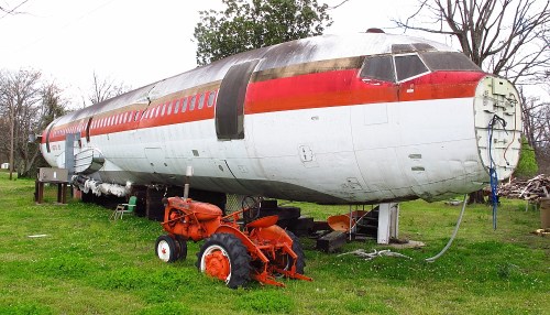 An old airplane fuselage sits on a platform in a grassy area, next to a vintage orange tractor.