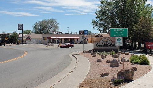 A road curves into Alamosa, Colorado, with a welcome sign and a small commercial area in the background.