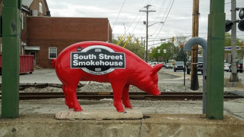 A red pig statue with a sign reading "South Street Smokehouse" near train tracks in a small town setting.