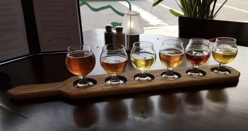 A wooden flight board with six small glasses of different colored beers on a table.