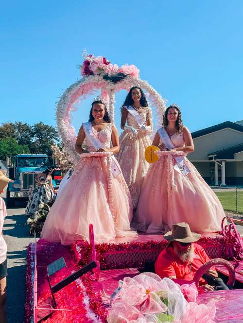 Three women in elegant pink gowns and tiaras smile from a decorated float during a parade under a clear blue sky.