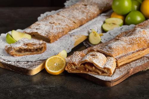 Two apple strudels dusted with powdered sugar, surrounded by fresh apples and lemons on wooden boards.