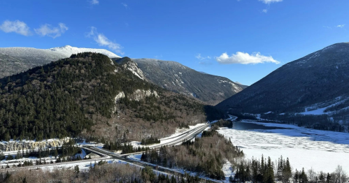 A scenic view of snow-covered mountains, a frozen lake, and a winding road under a clear blue sky.