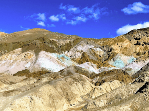 A colorful mountainous landscape with layers of earth tones under a bright blue sky and scattered clouds.