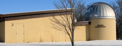 A yellow observatory building with a silver dome, surrounded by snow and bare trees under a clear blue sky.