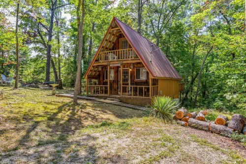 A charming A-frame cabin surrounded by trees, with a red roof and wooden porch, set in a lush green landscape.