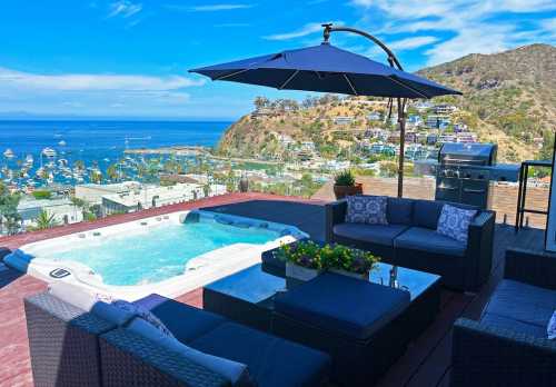 A scenic patio with a hot tub, seating area, and an umbrella, overlooking a coastal view and marina.