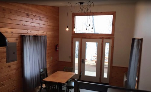 Cozy interior with wooden walls, a dining table, and large windows overlooking a lake. Soft lighting adds warmth.