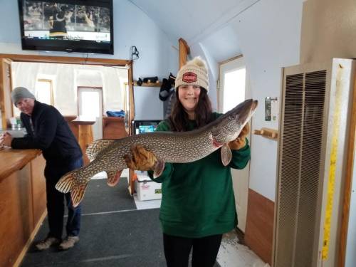 A person in a green sweater holds a large pike fish inside an ice fishing hut, with a TV in the background.